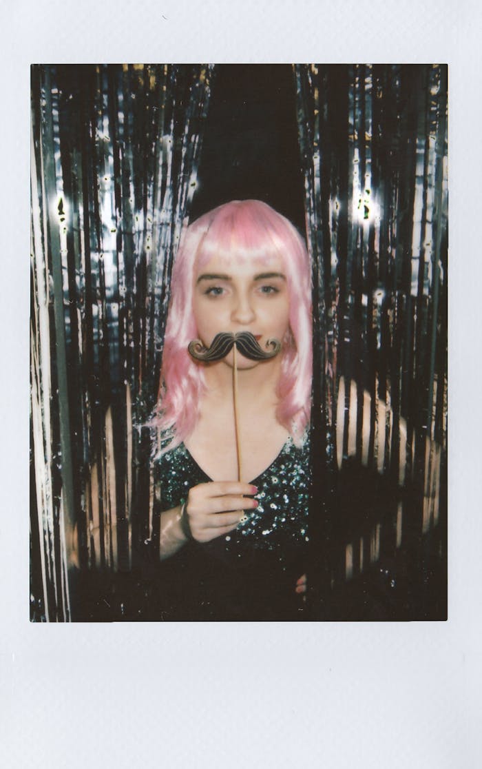 Home A woman with pink hair poses with a novelty mustache in a sequin top, creating a playful vibe.